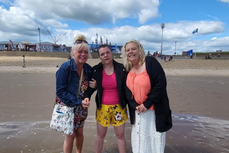 Photo of care home resident with two carers stood on a beach